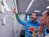man wearing blue Windows sweater holding sticky note on white board sales team crm software happy office charts