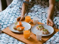 Wellness Challenges That Can Improve Your Life In Just 30 Days person serving pastries on white ceramic plates with fruit juice glasses on wooden tray on top of bed morning routine, healthy habits, casual home setup