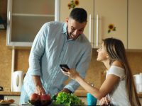 Couple looking at phone in a bright kitchen couple budgeting together, smartphone budget app, home kitchen table